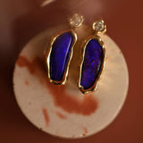 Gold earrings with blue opals on a round white card against a brown background