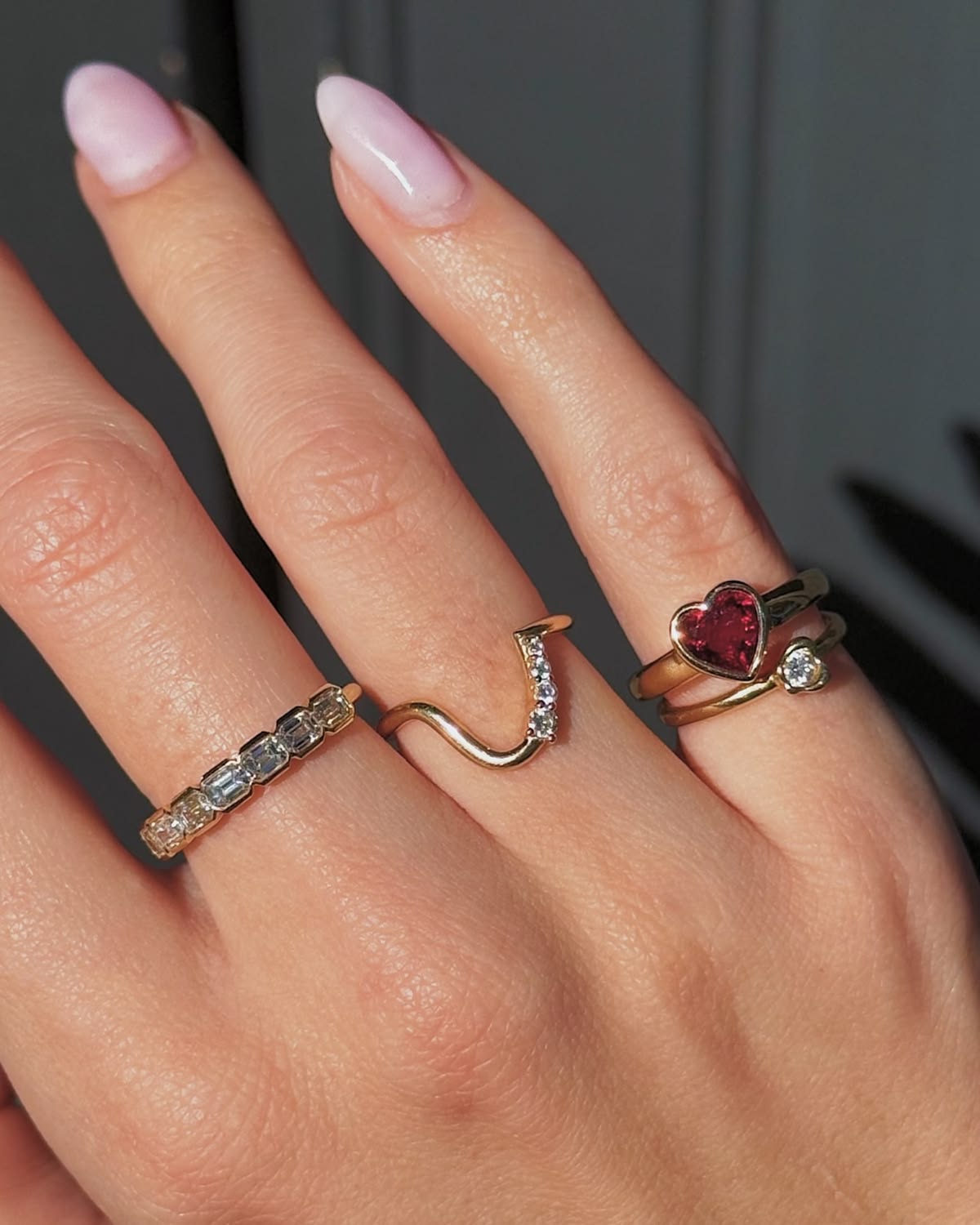 A close-up of a hand wearing three gold rings: a silver-and-gold pavé band ring, a wavy gold ring with small clear stones, and a gold ring with a heart-shaped red gemstone and a small clear accent stone.