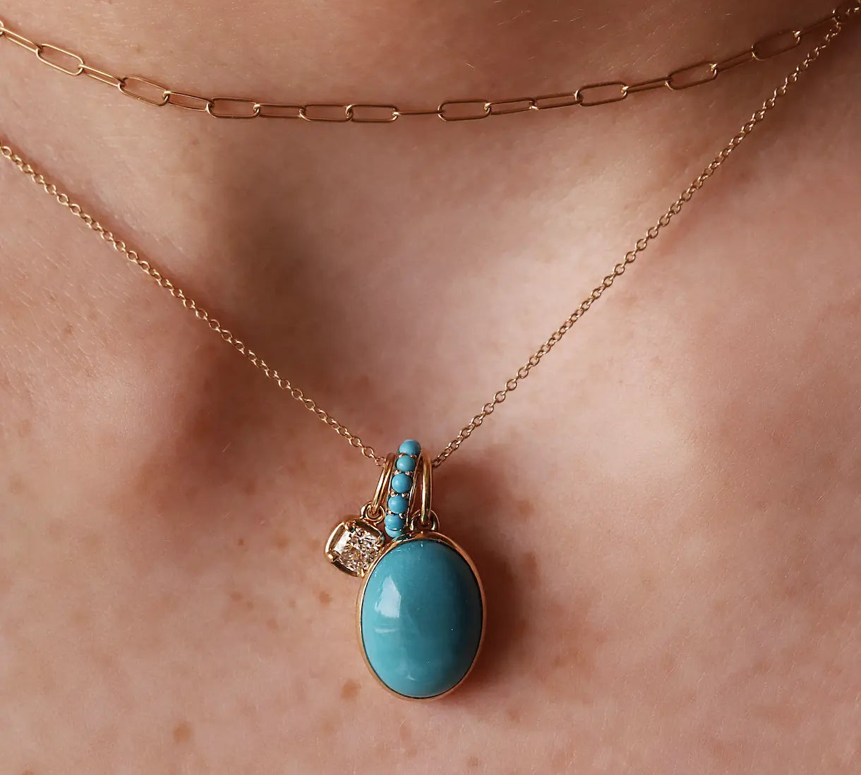 A close-up of a person's neck wearing a rose gold layered necklace set, featuring a delicate chain and a longer paperclip chain with a pendant. The pendant has a large oval light blue stone in a yellow gold setting, with a small gold charm and tiny clear stones.