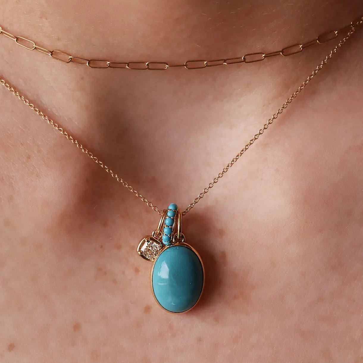 A close-up of a person's neck wearing a rose gold layered necklace set, featuring a delicate chain and a longer paperclip chain with a pendant. The pendant has a large oval light blue stone in a yellow gold setting, with a small gold charm and tiny clear stones.
