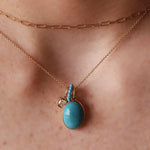 A close-up of a person's neck wearing a rose gold layered necklace set, featuring a delicate chain and a longer paperclip chain with a pendant. The pendant has a large oval light blue stone in a yellow gold setting, with a small gold charm and tiny clear stones.