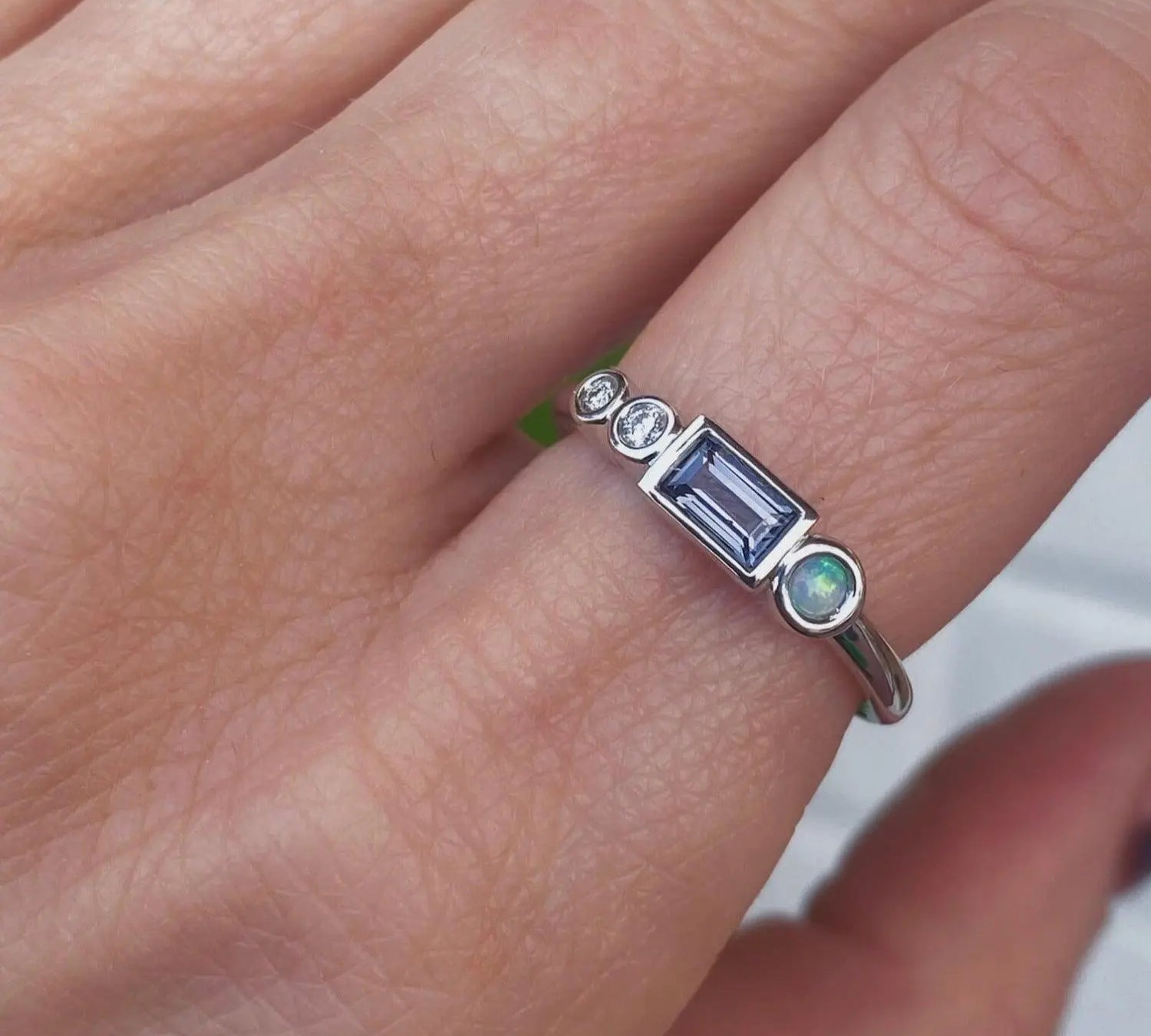Image of a marrow fine linear band on a pointer finger. The one of a kind ring has diamonds, a spinel baguette, and a small opal round. All stones are bezel set and in 14K white gold.