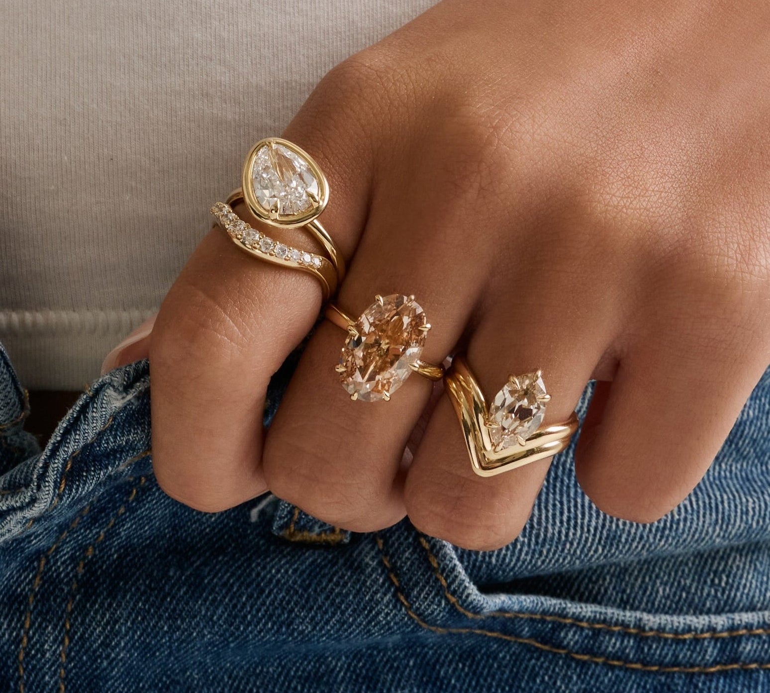 A close-up lifestyle shot of a person wearing multiple gold rings with clear gemstones on their hands, styled with a beige ribbed top and blue jeans.