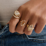 A close-up lifestyle shot of a person wearing multiple gold rings with clear gemstones on their hands, styled with a beige ribbed top and blue jeans.
