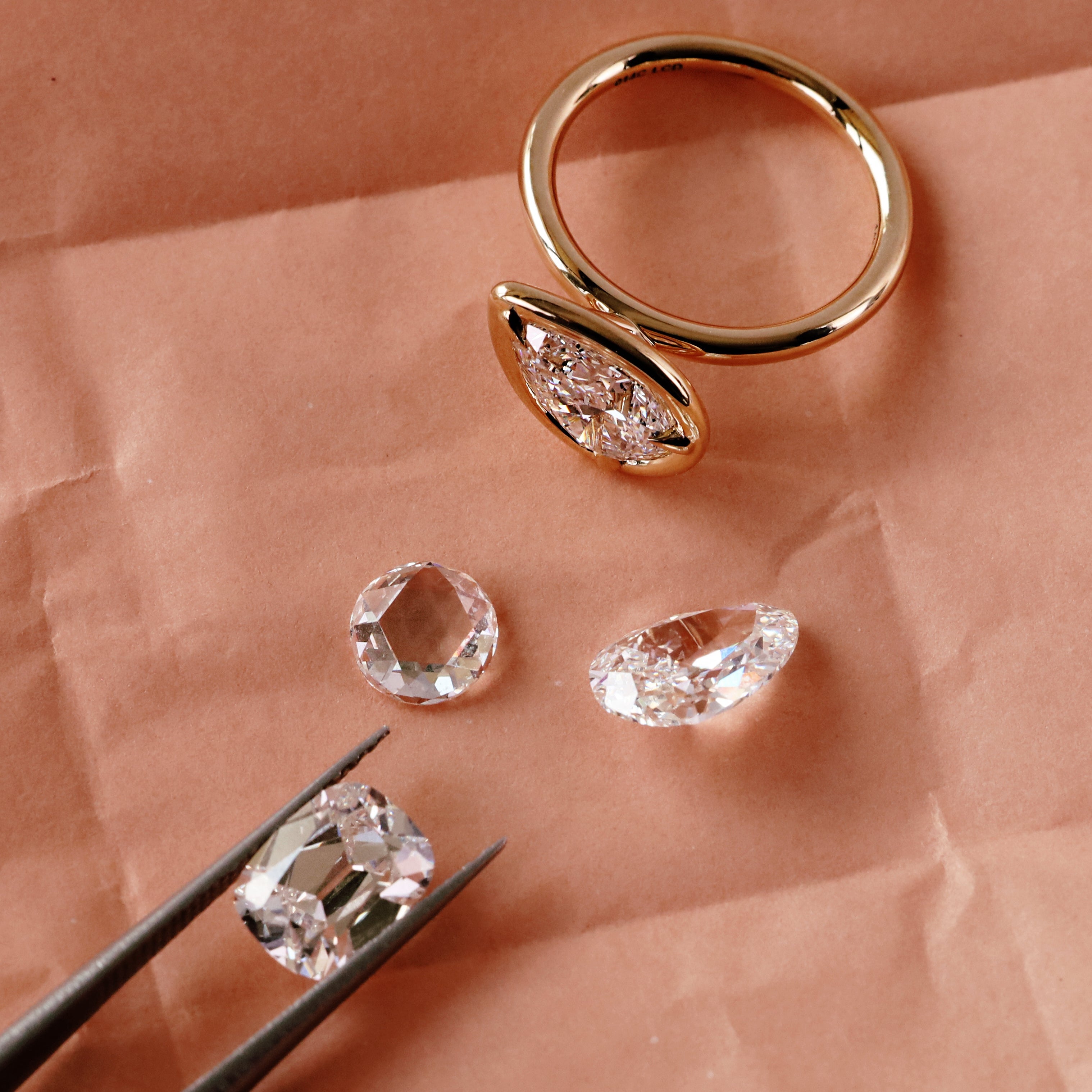 A gold ring with a marquise-shaped clear gemstone setting, displayed on peach-colored paper alongside three loose clear gemstones and black tweezers holding one stone.
