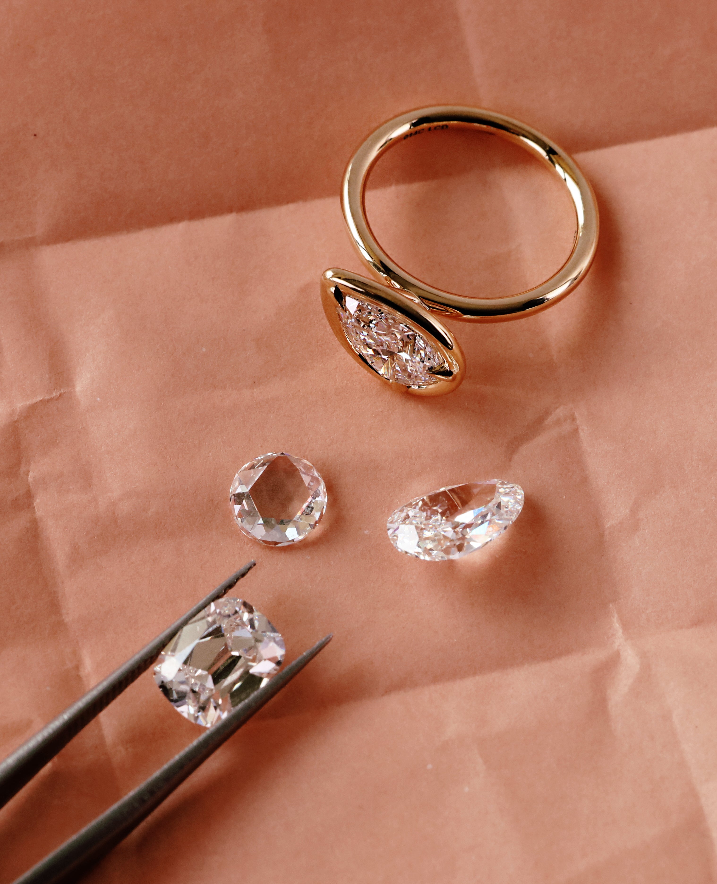 A gold ring with a marquise-shaped clear gemstone setting, displayed on peach-colored paper alongside three loose clear gemstones and black tweezers holding one stone.