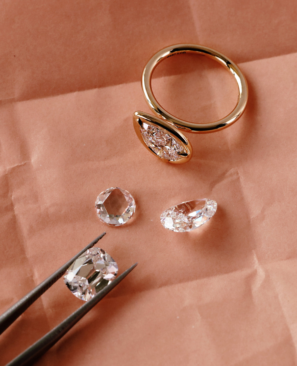 A gold ring with a marquise-shaped clear gemstone setting, displayed on peach-colored paper alongside three loose clear gemstones and black tweezers holding one stone.