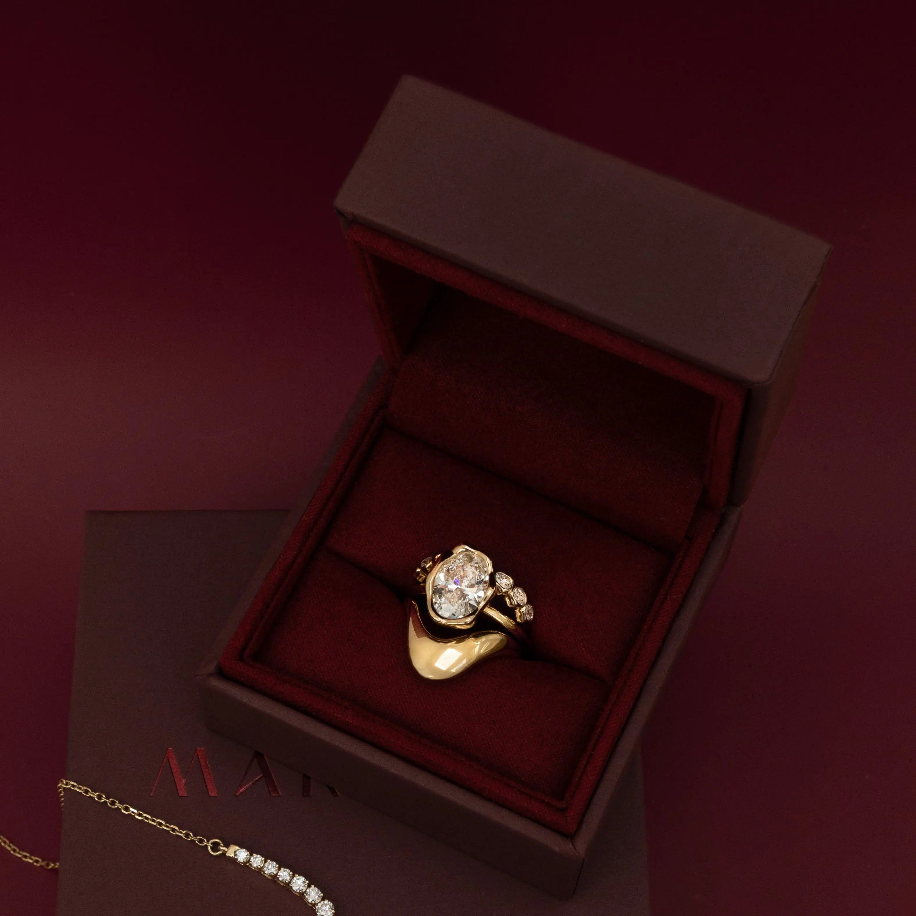 The Abstract Bezel Oval Engagement Ring stack in an open burgundy velvet ring box with a diamond necklace draped beside it