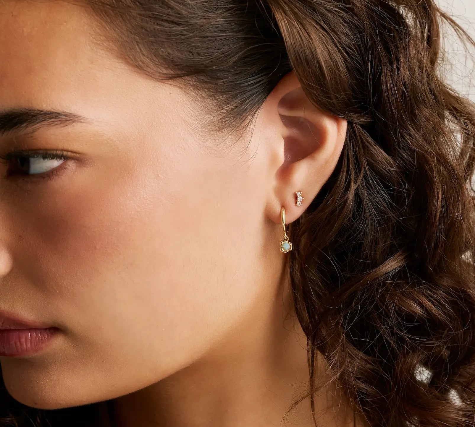 close up of woman's side profile with brown curly hair wearing opal drop hoops and a 3 diamond stud on second piercing.