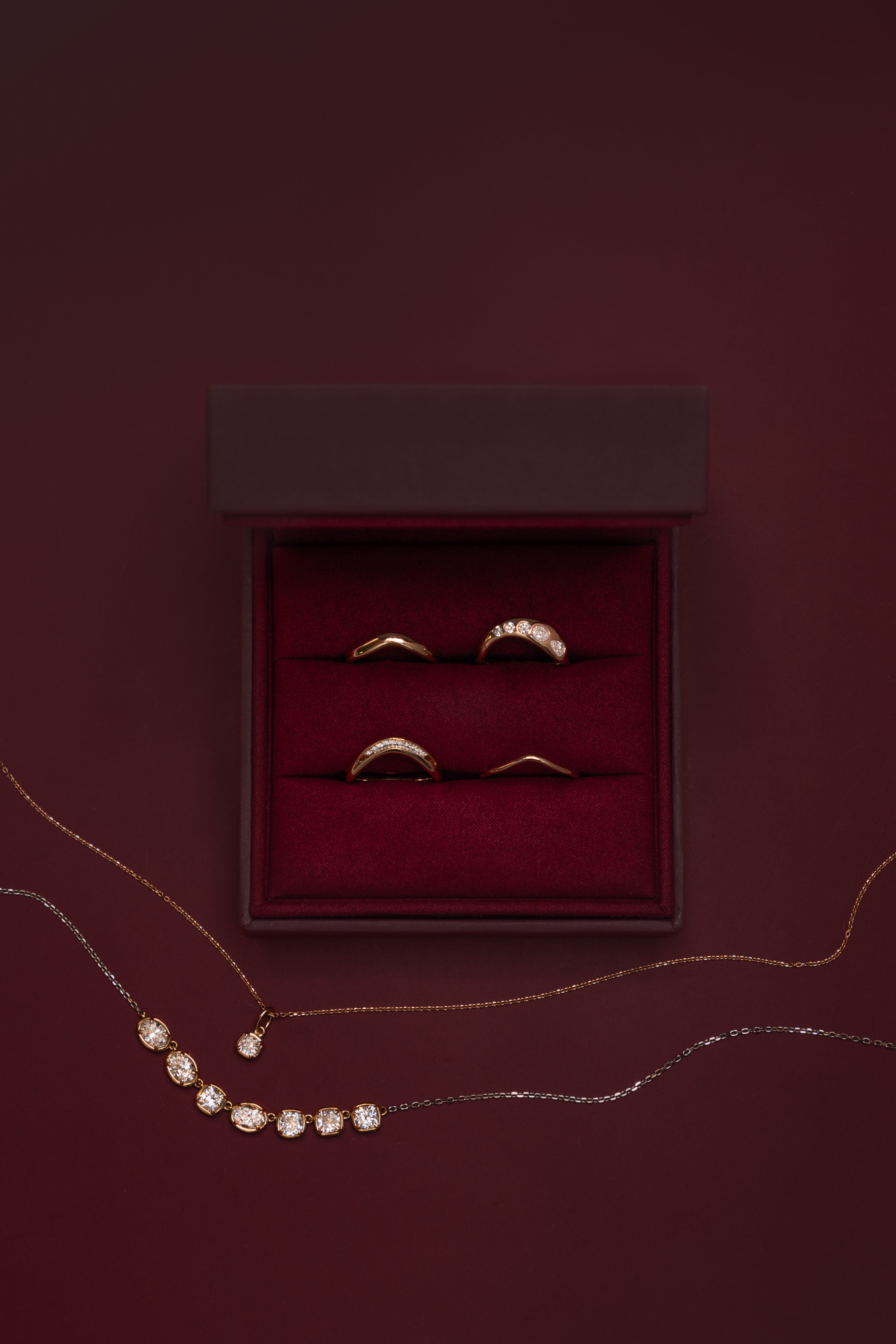 Marrow Fine wave stacking rings in an open burgundy velvet ring box: the White Diamond Estelle Wave Band, White Diamond Lola Bombe Wave Band and Everyday Wave Bands in 3mm and 1.5mm, with the Lisse Diamond Cushion Charm and Lisse Seven Stone Diamond Necklace draped below