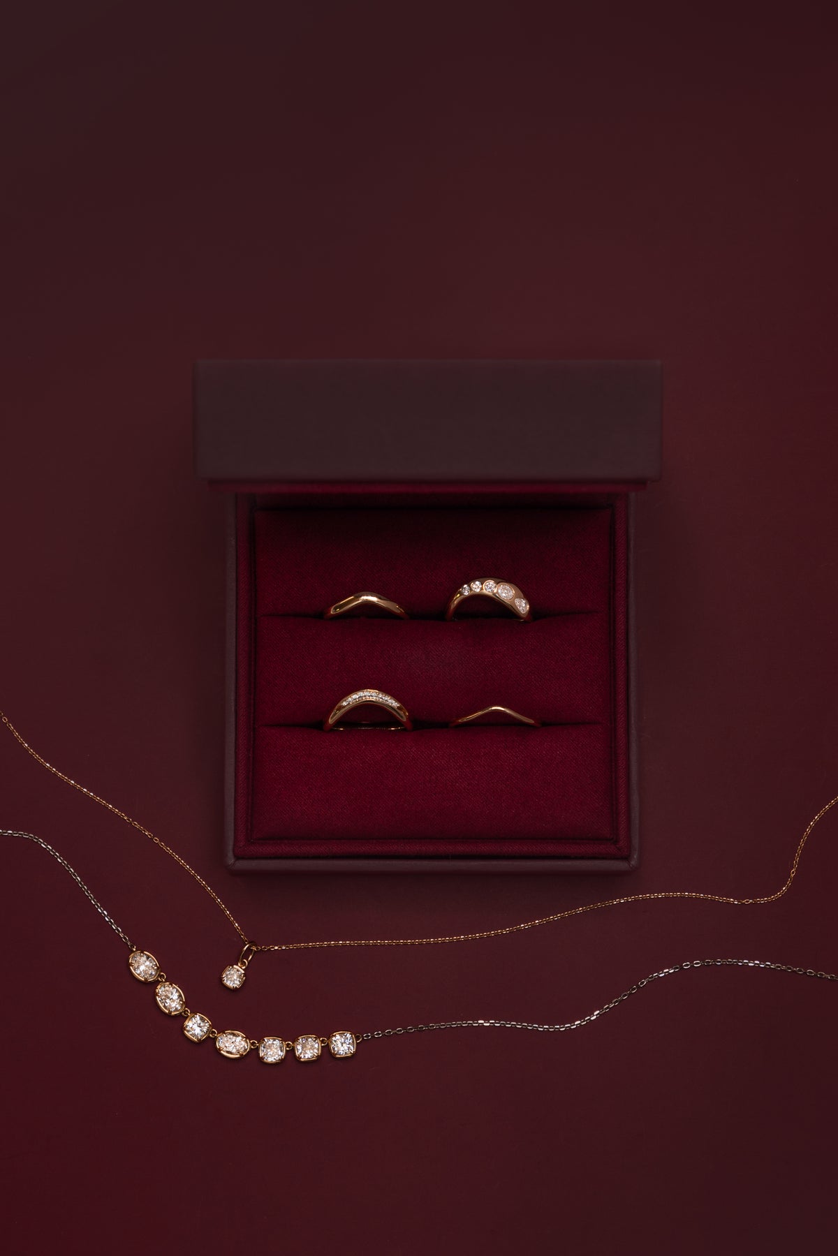 Marrow Fine wave stacking rings in an open burgundy velvet ring box: the White Diamond Estelle Wave Band, White Diamond Lola Bombe Wave Band and Everyday Wave Bands in 3mm and 1.5mm, with the Lisse Diamond Cushion Charm and Lisse Seven Stone Diamond Necklace draped below