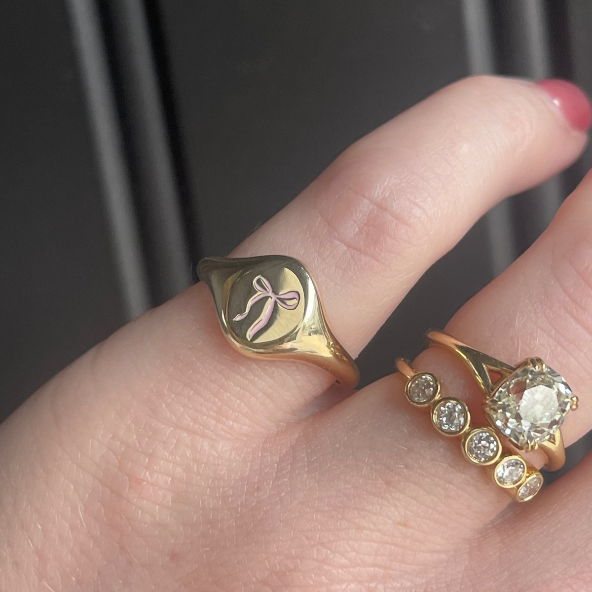 A close-up of a hand wearing two gold rings. The primary ring is a signet-style ring with a polished gold leaf finish and an engraved bow motif.