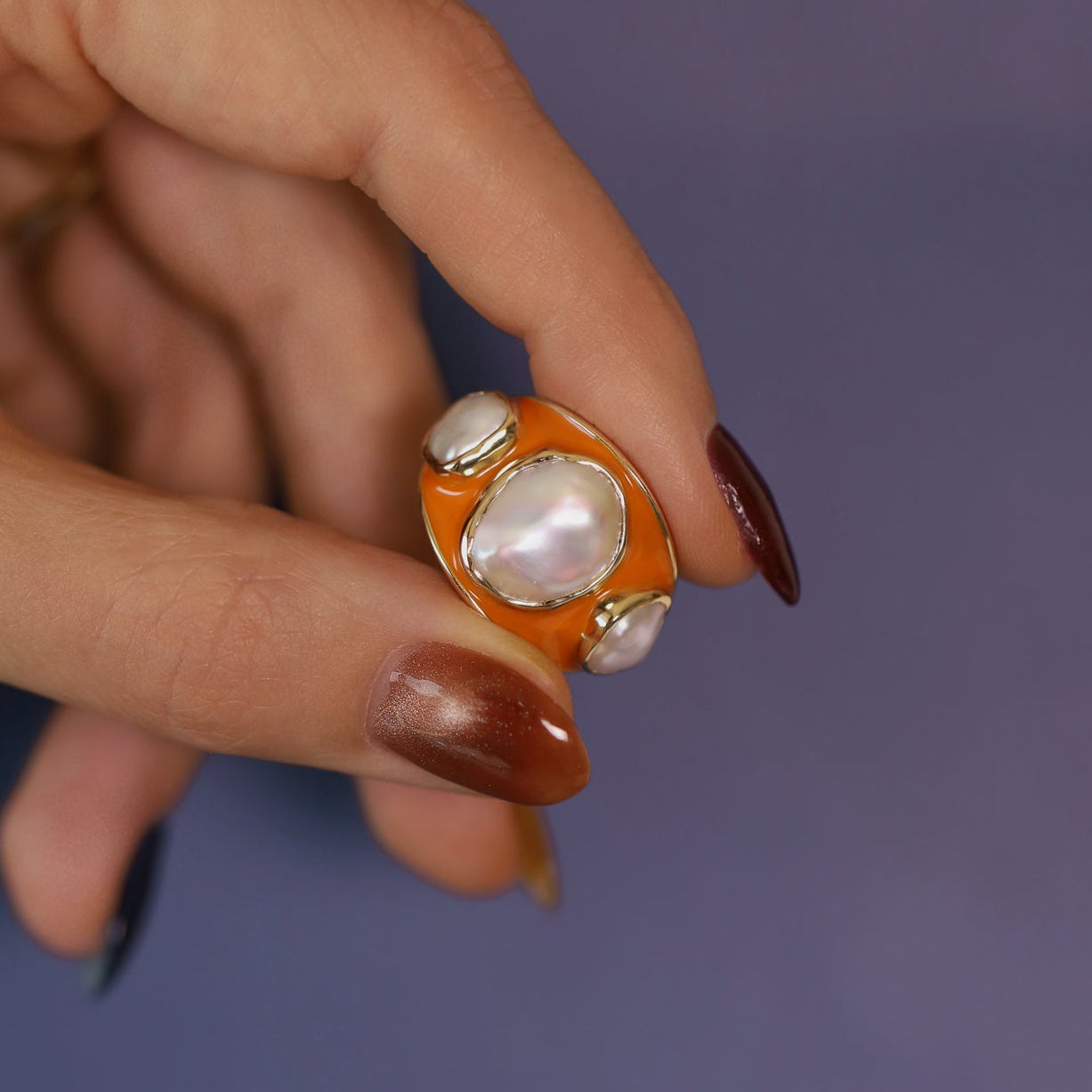 Hand holding the Baroque Keshi Pearl and Burnt Orange Enamel Ring against a purple background