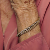 Close-up of an elderly person's wrist wearing a gold bracelet with clear stones on a pink shirt background.