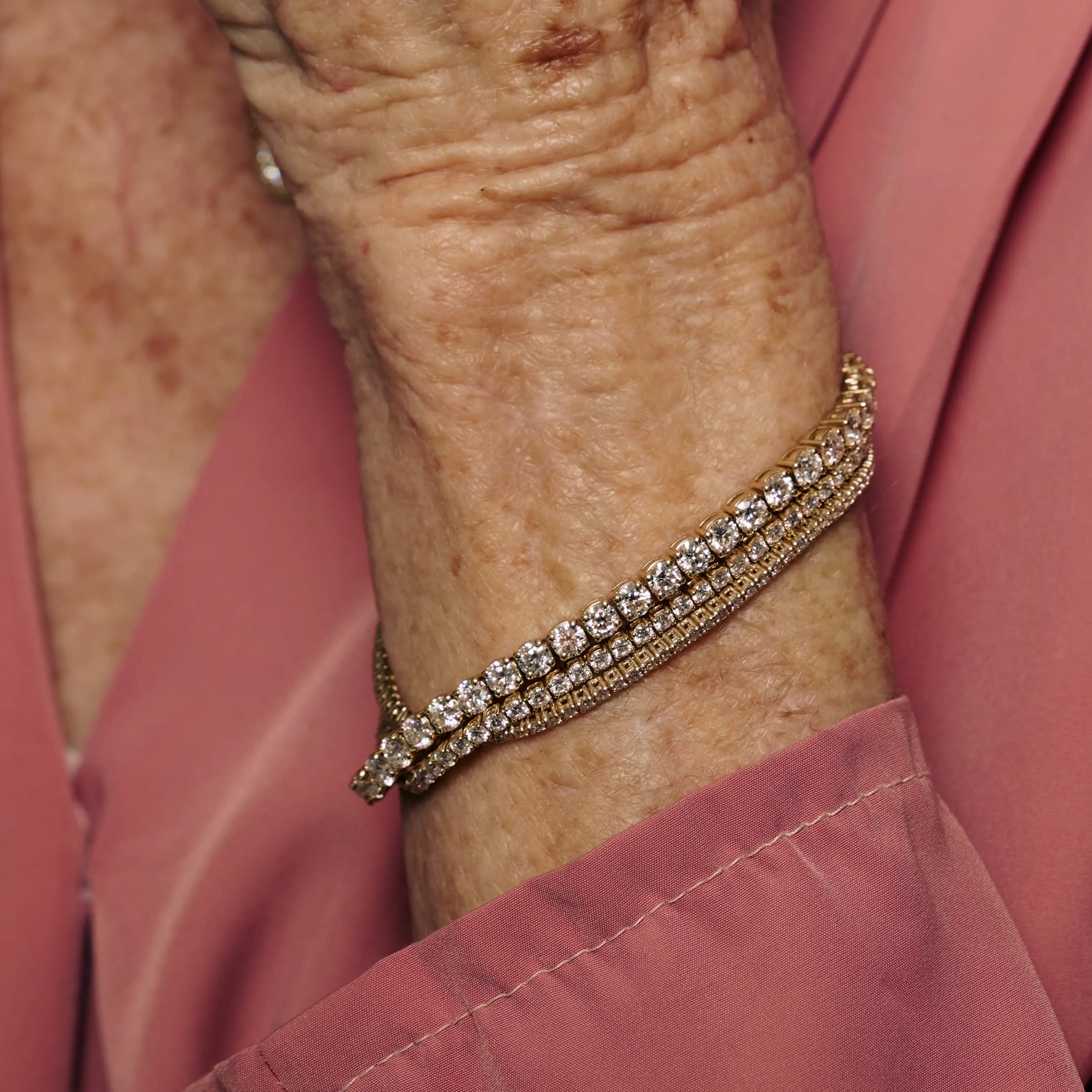 Close-up of an elderly person's wrist wearing a gold bracelet with clear stones on a pink shirt background.