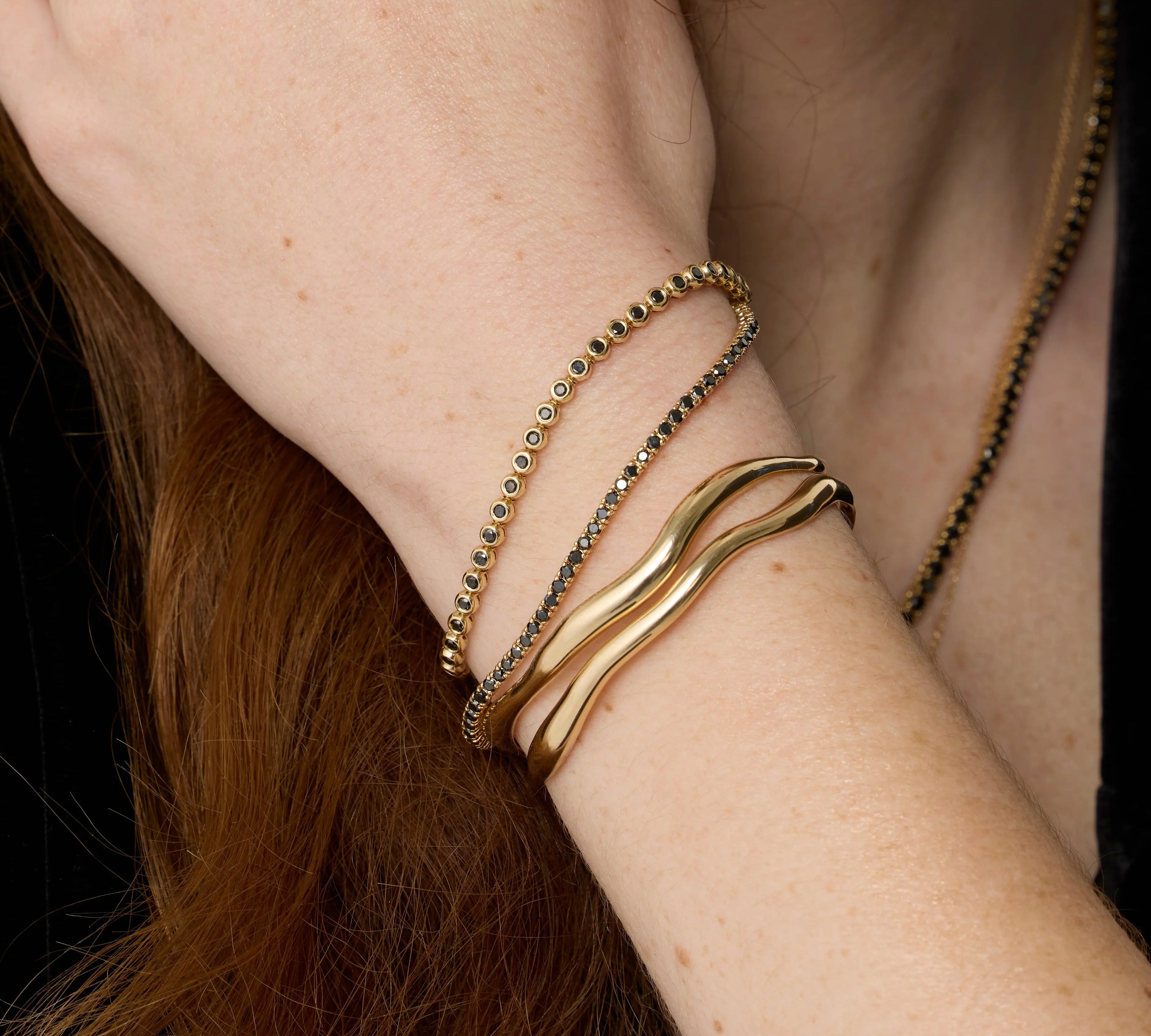 Close up of Curved Freeform Gold Cuff + Black Diamond Tennis Bracelet + Black Diamond Bezeled Tennis Bracelet on wrist
