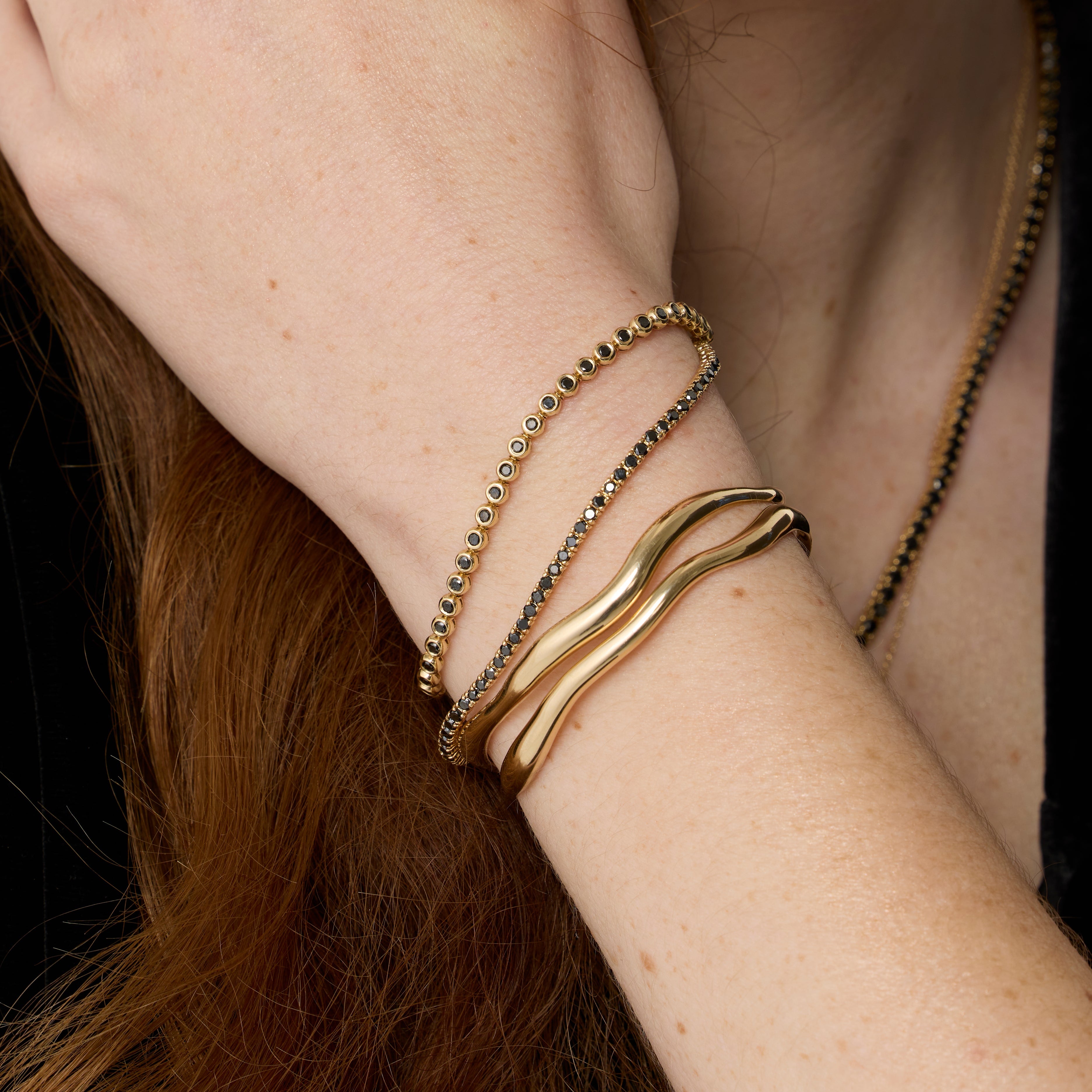 Close up of Curved Freeform Gold Cuff + Black Diamond Tennis Bracelet + Black Diamond Bezeled Tennis Bracelet on wrist