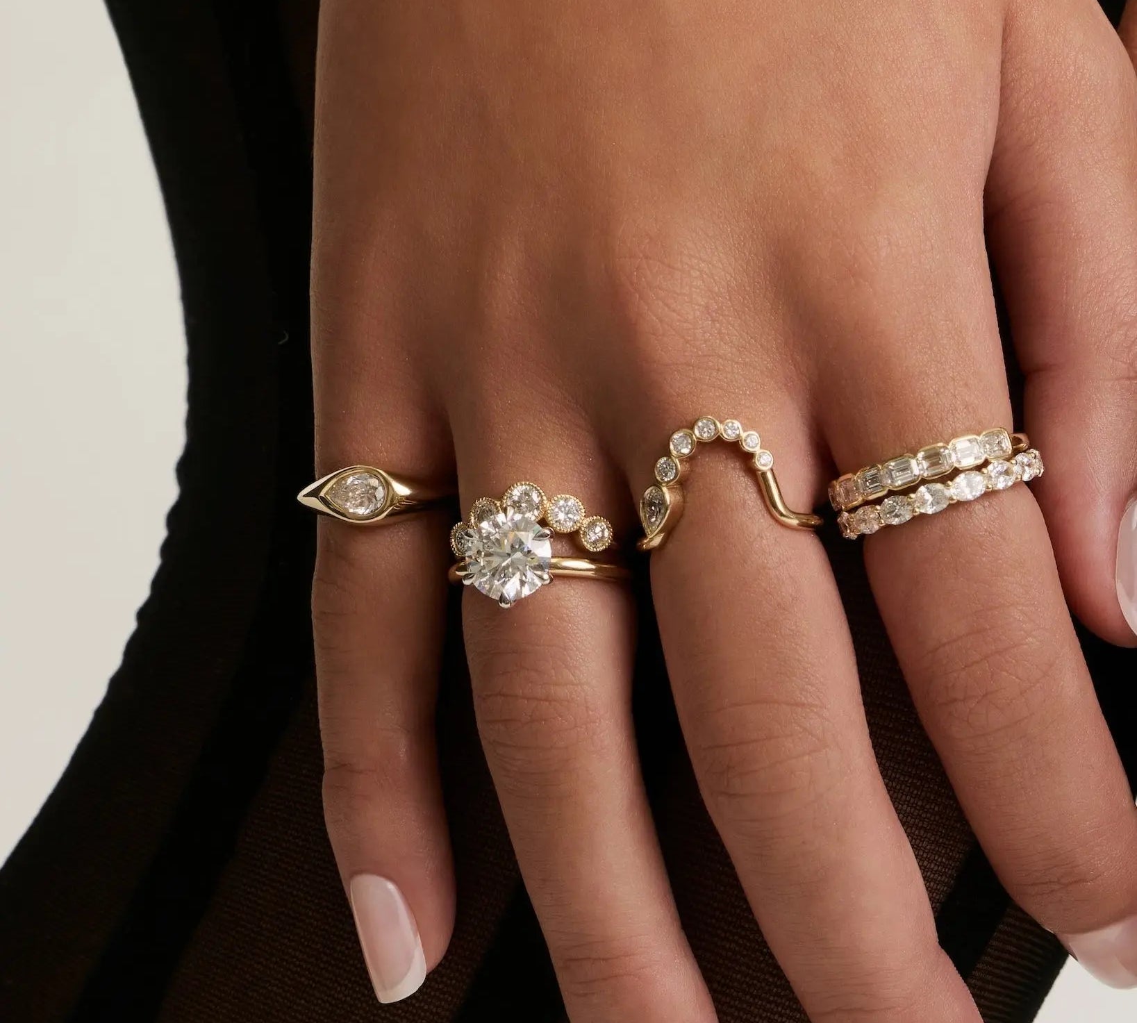 A close-up of a person's hands and legs wearing multiple gold rings and bracelets, styled with sheer black tights against a light neutral background.