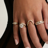 A close-up of a person's hands and legs wearing multiple gold rings and bracelets, styled with sheer black tights against a light neutral background.