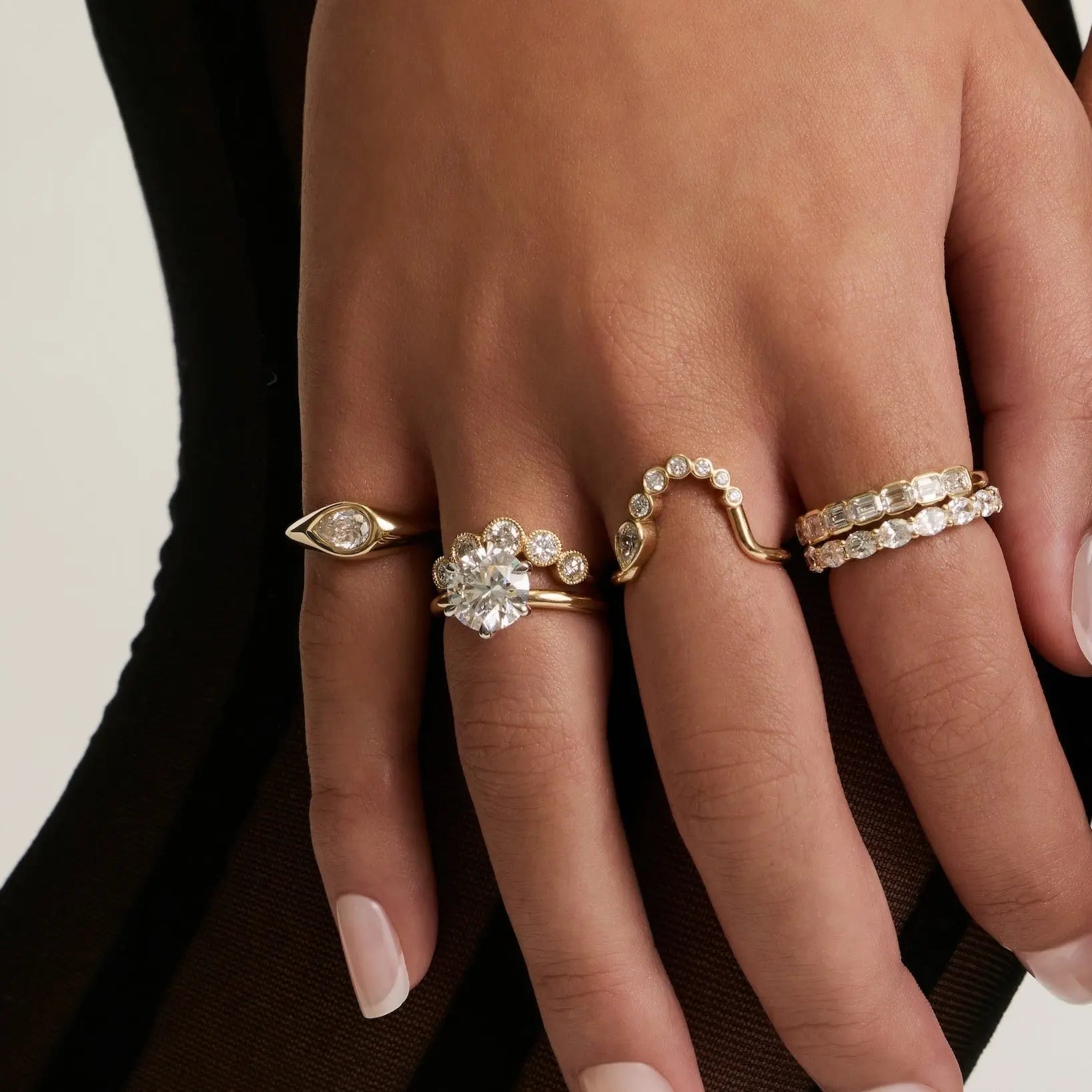 A close-up of a person's hands and legs wearing multiple gold rings and bracelets, styled with sheer black tights against a light neutral background.