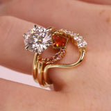 A gold ring with a large clear round-cut center stone, a square orange gemstone, and curved rows of small clear and pink stones, shown worn on a hand.