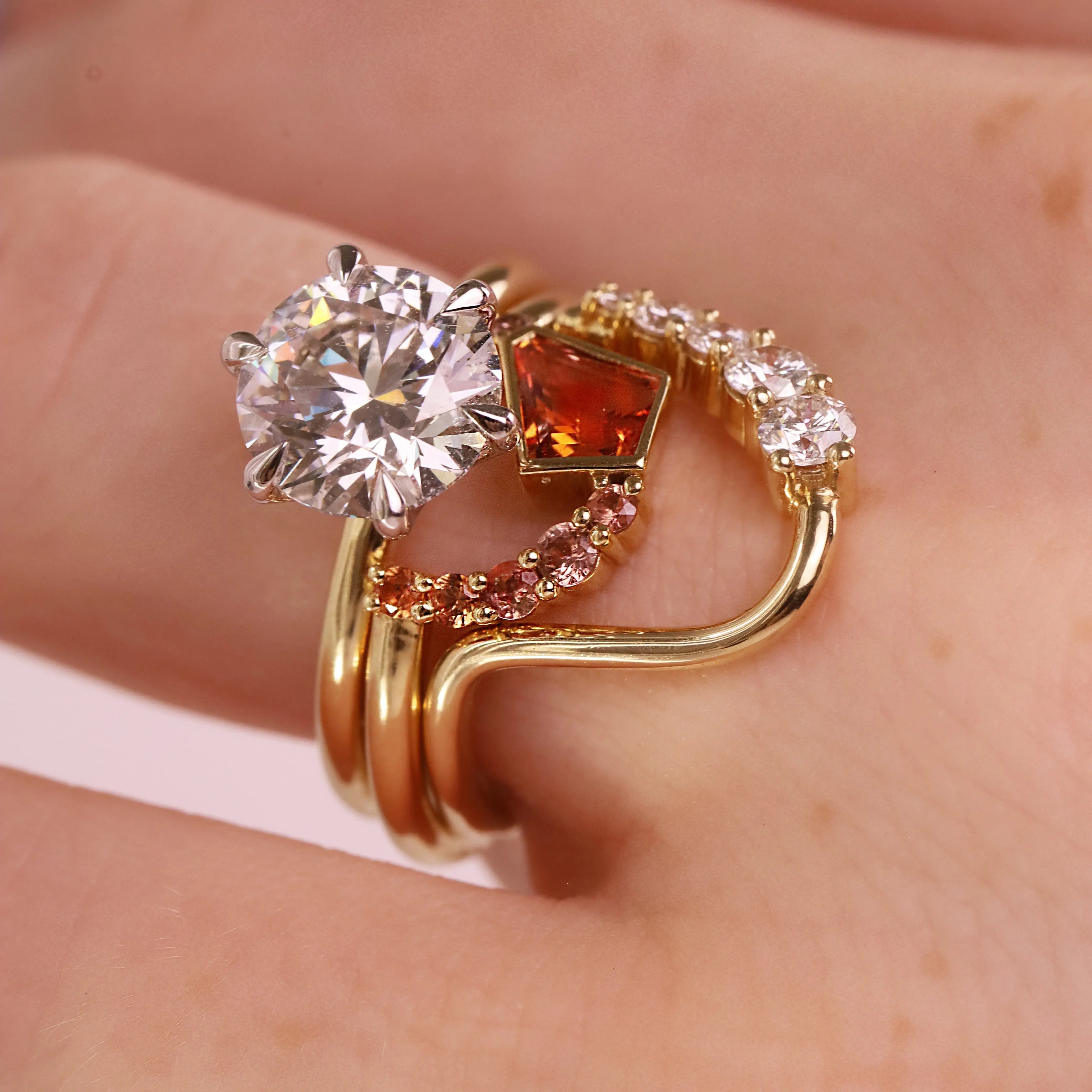 A gold ring with a large clear round-cut center stone, a square orange gemstone, and curved rows of small clear and pink stones, shown worn on a hand.