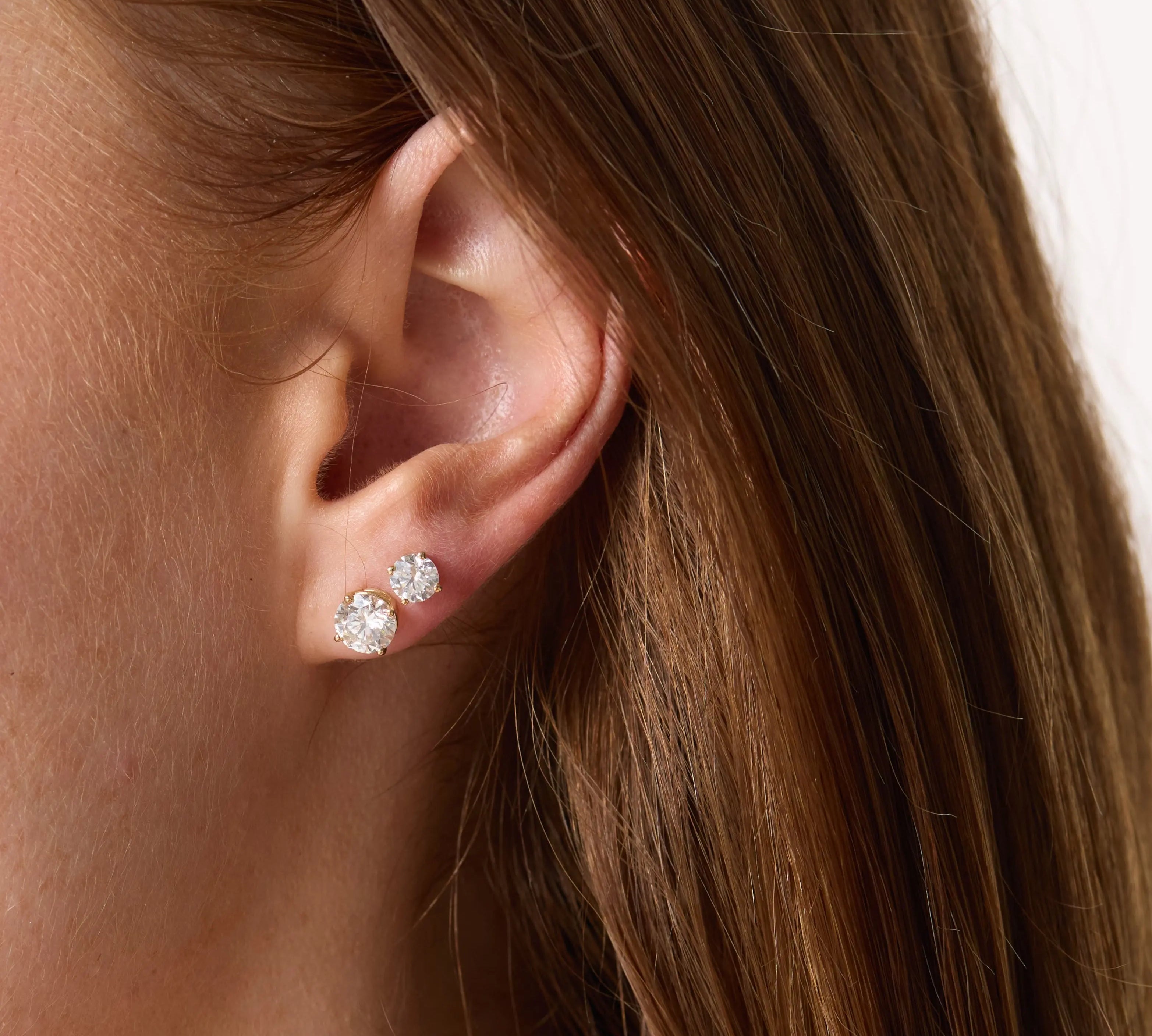 Close-up of a person wearing two diamond stud earrings with a neutral background.