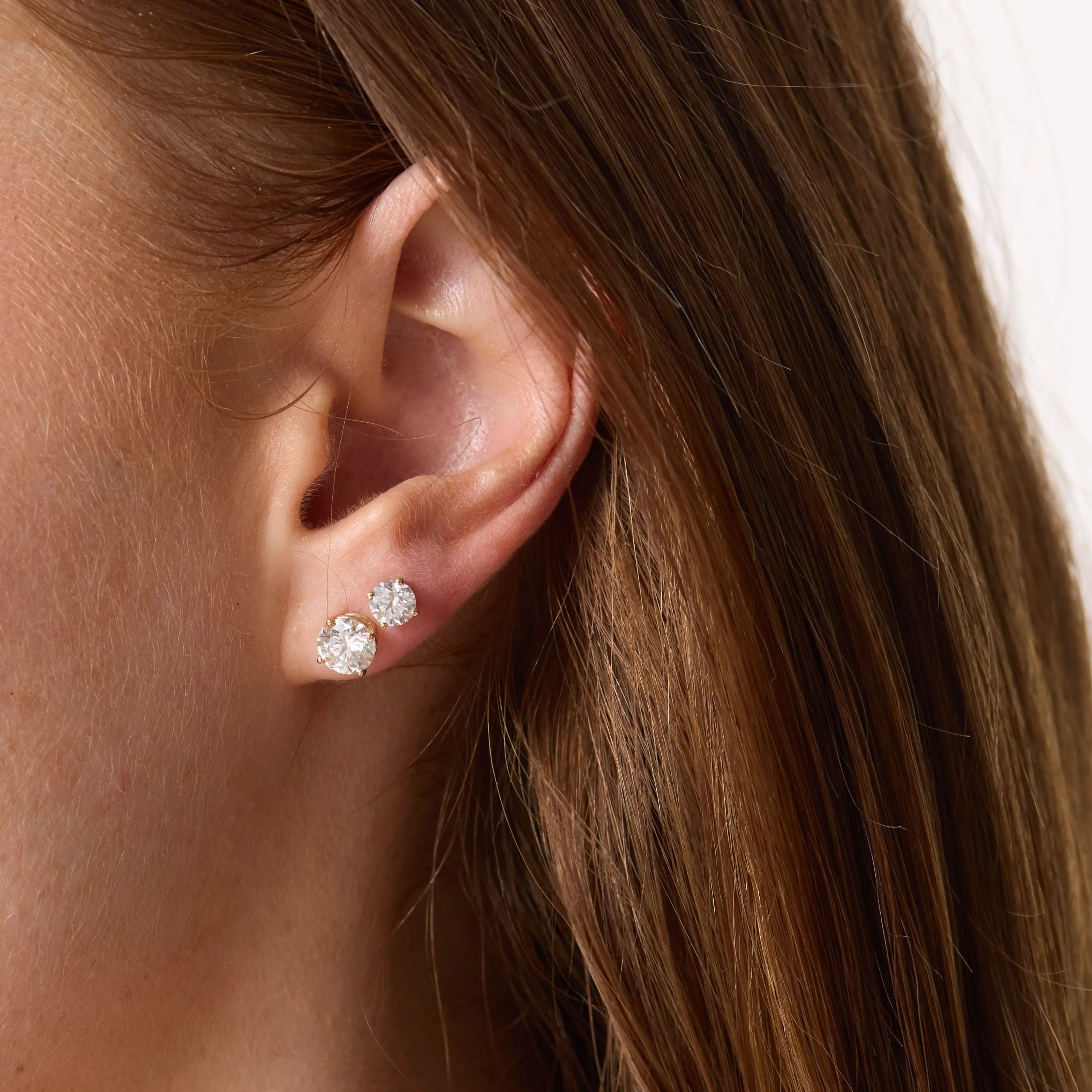 Close-up of a person wearing two diamond stud earrings with a neutral background.