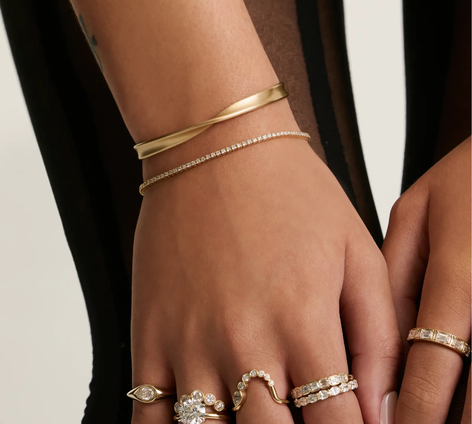 A close-up of a person's hand and wrist wearing multiple delicate rings and bracelets. The jewelry includes gold bands, a thin diamond-studded bracelet, and several rings with clear gemstones set in white metal, shown against a dark and light neutral background.