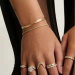 A close-up of a person's hand and wrist wearing multiple delicate rings and bracelets. The jewelry includes gold bands, a thin diamond-studded bracelet, and several rings with clear gemstones set in white metal, shown against a dark and light neutral background.