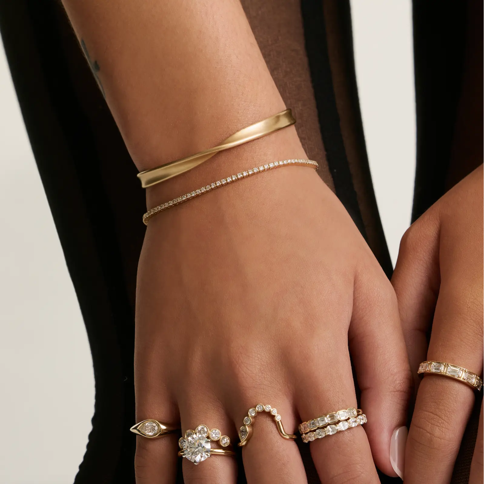A close-up of a person's hand and wrist wearing multiple delicate rings and bracelets. The jewelry includes gold bands, a thin diamond-studded bracelet, and several rings with clear gemstones set in white metal, shown against a dark and light neutral background.