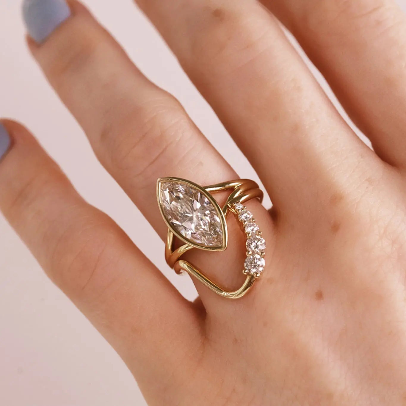 A gold ring with a marquise-cut clear diamond on split shank. Stacked with a wavy diamond band. Shown worn on a hand.