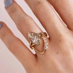 A gold ring with a marquise-cut clear diamond on split shank. Stacked with a wavy diamond band. Shown worn on a hand.