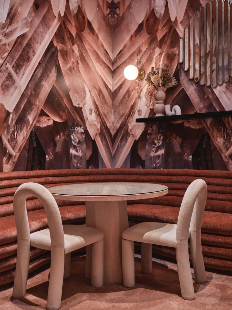A round dining table with a glass top and curved cream-colored chairs, set in a uniquely designed room with a crystal-like pink and brown wall and a terracotta-colored curved bench.