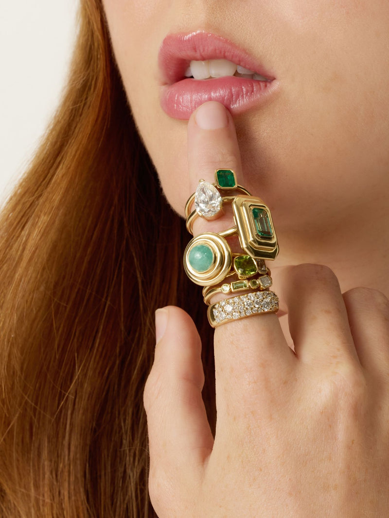 A close-up of a person's hand and lips wearing a stack of gold rings with green and clear gemstones, against a light neutral background.