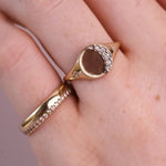 A close-up of a hand wearing two gold rings. The larger ring features a semi-circular face with a row of small clear stones on one side and a smooth brown center.
