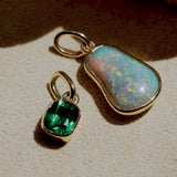 Two gold pendants with a green gemstone and an opal-like stone on a beige surface.