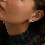 Close-up of a woman's ear wearing a pair of gold and white gemstone stud earrings, with her face and neck visible against a soft neutral background.