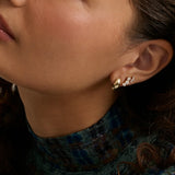 Close-up of a woman's ear wearing a pair of gold and white gemstone stud earrings, with her face and neck visible against a soft neutral background.
