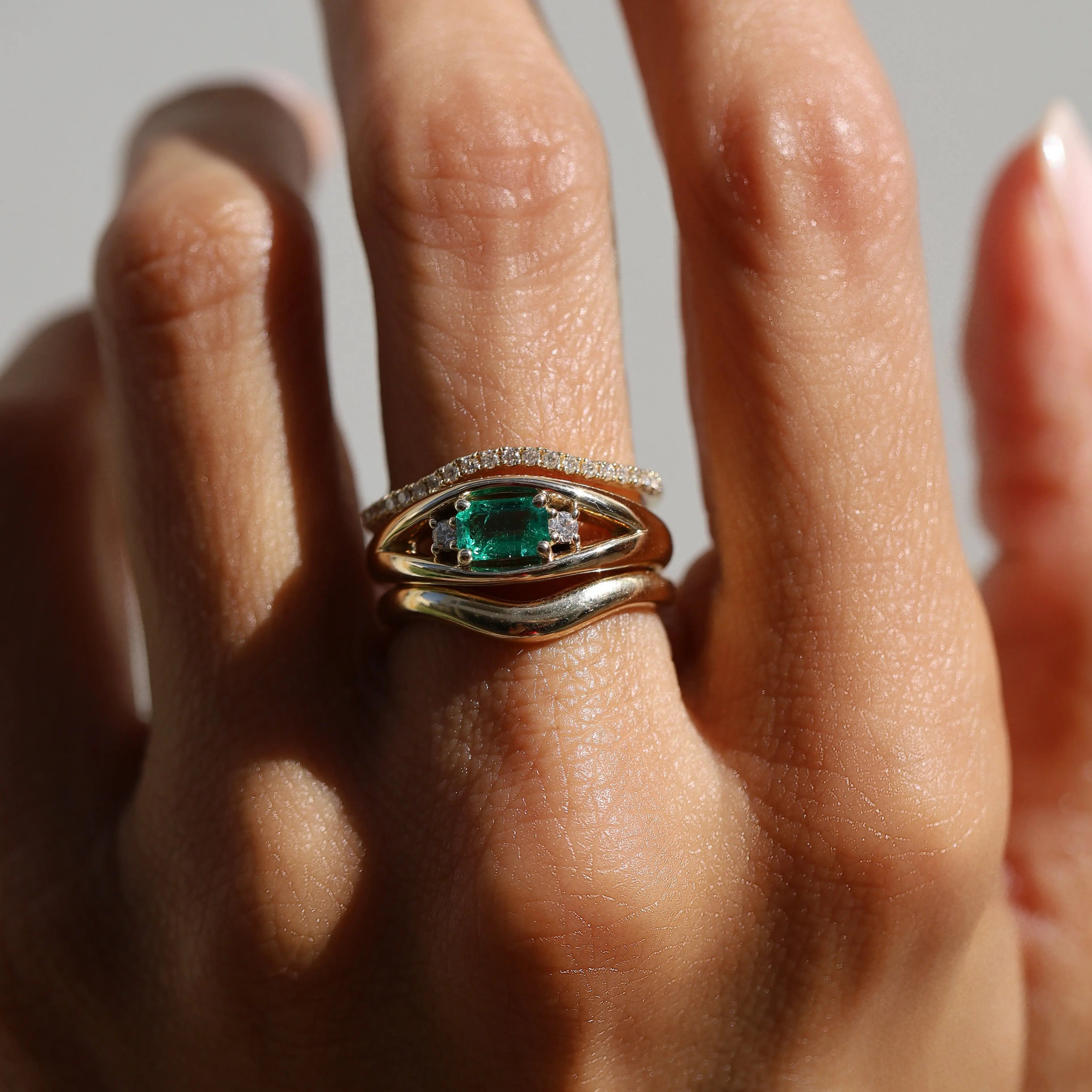 Gold ring with emerald and diamonds on a finger against a neutral background