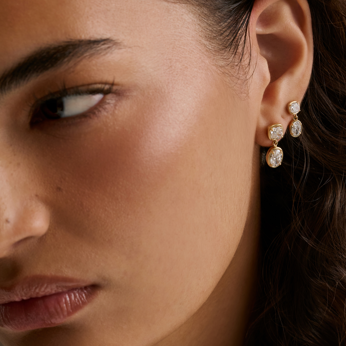 A close-up of a model's ear wearing a three-stone gold and diamond drop earring with diamonds.