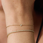 Two delicate gold bracelets worn on a person's wrist, including a fine chain with a single round clear stone and a tennis-style bracelet with multiple small clear stones.