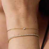 Two delicate gold bracelets worn on a person's wrist, including a fine chain with a single round clear stone and a tennis-style bracelet with multiple small clear stones.