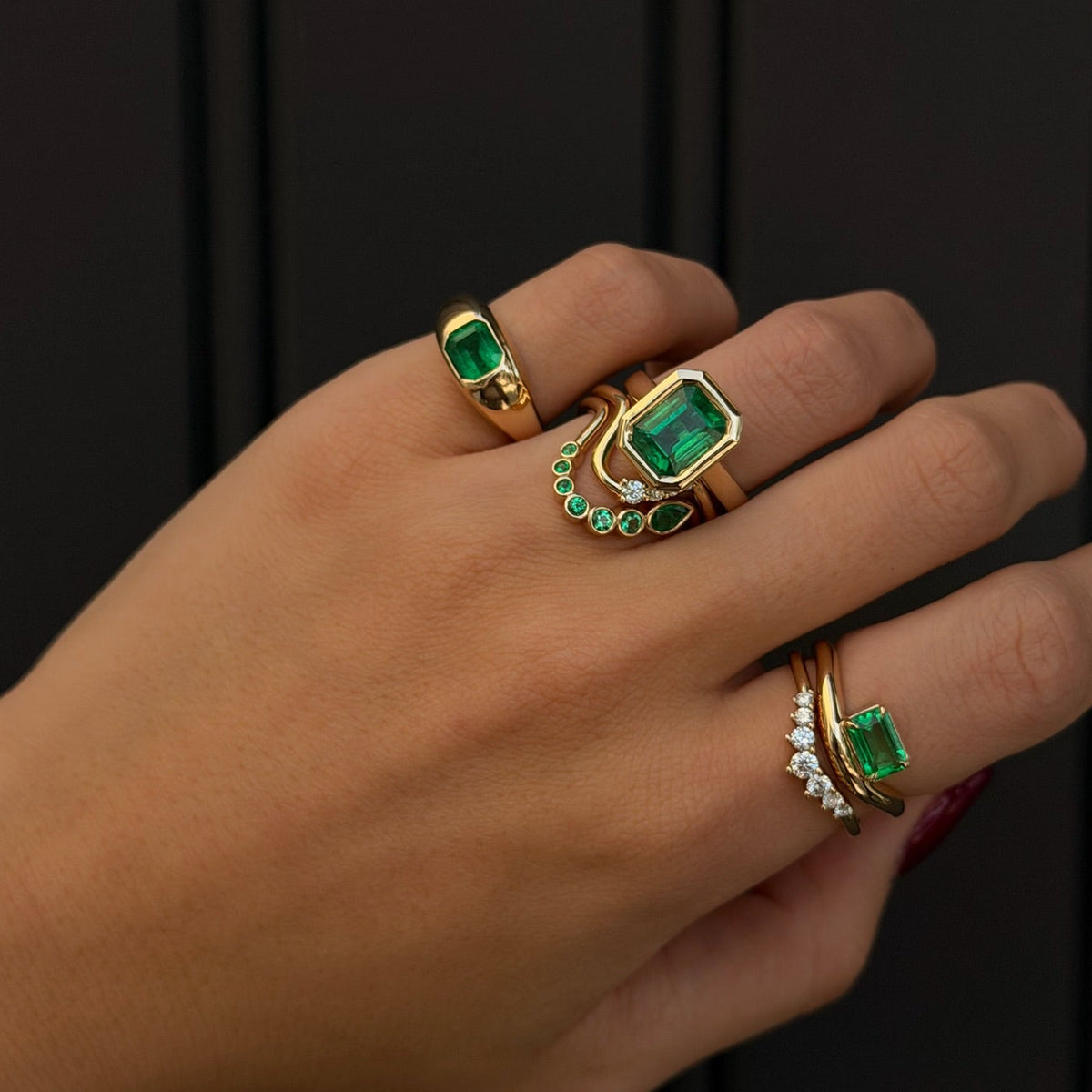 A close-up of a hand wearing four gold rings with emerald-cut green stones and small clear accent stones, set against a dark paneled background.
