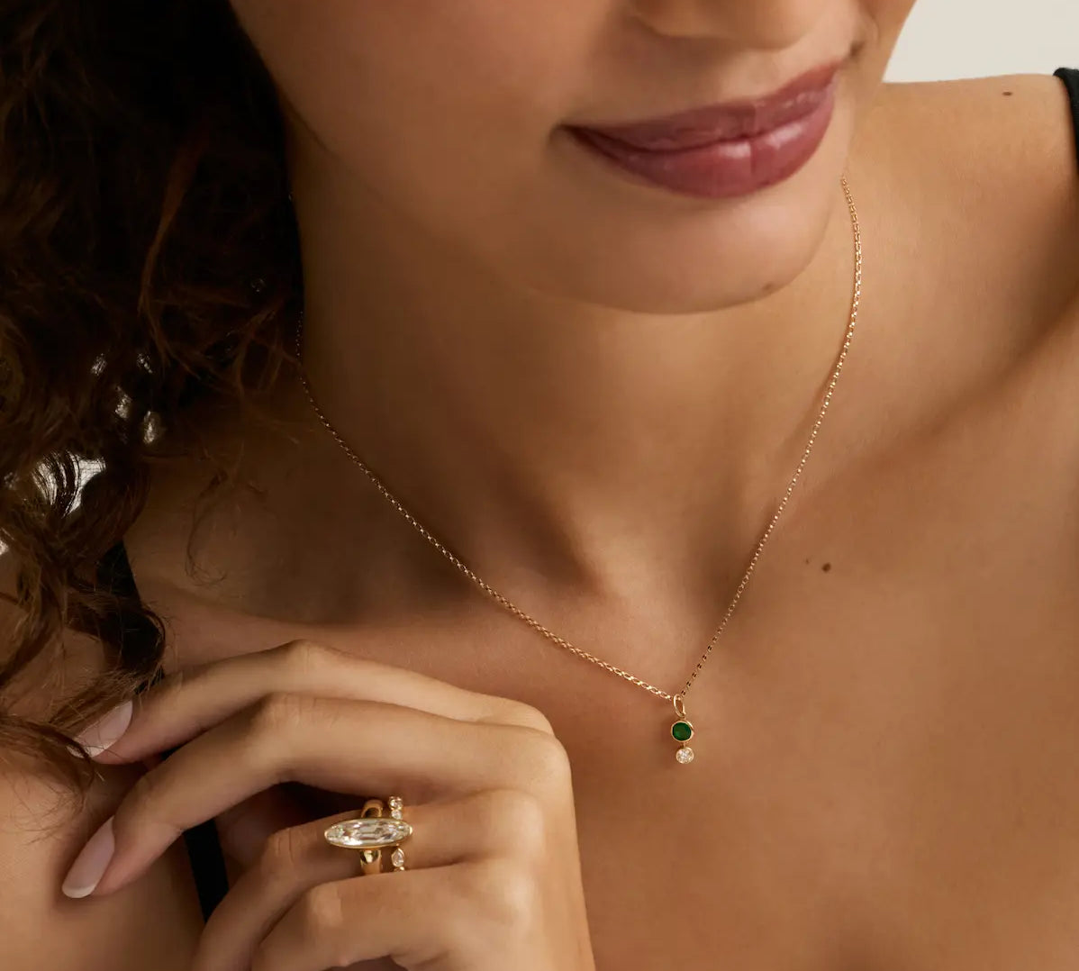 A close-up lifestyle shot of a woman wearing a delicate gold chain necklace with a small green and white bead pendant, and a gold ring on her finger.