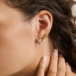 A close-up of a woman's ear wearing a stack of gold earrings, including a small hoop, a stud with a clear gemstone, and a dangling oval charm with a green accent stone.