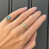 A close-up of a hand wearing two gold rings against a dark gray paneled background. One ring features a blue and white cameo-style stone, and the other is a delicate gold band with small clear stones.