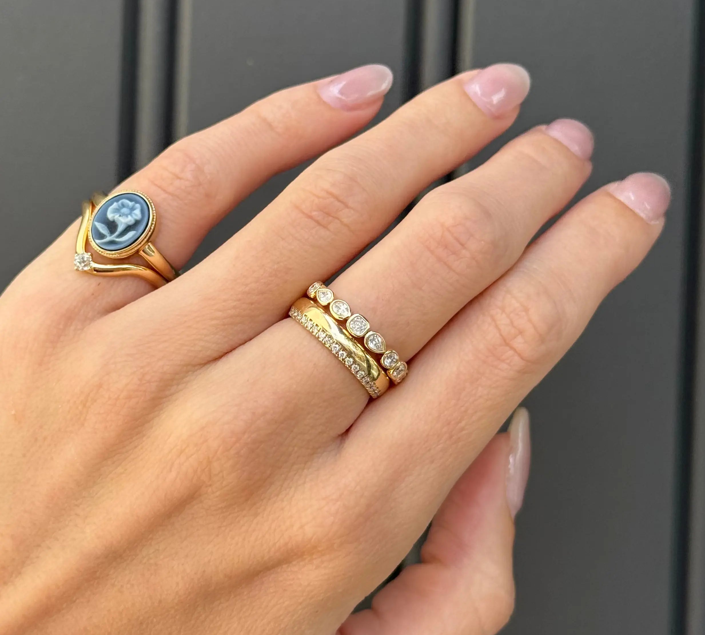 A close-up of a hand wearing two gold rings against a dark gray paneled background. One ring features a blue and white cameo-style stone, and the other is a delicate gold band with small clear stones.