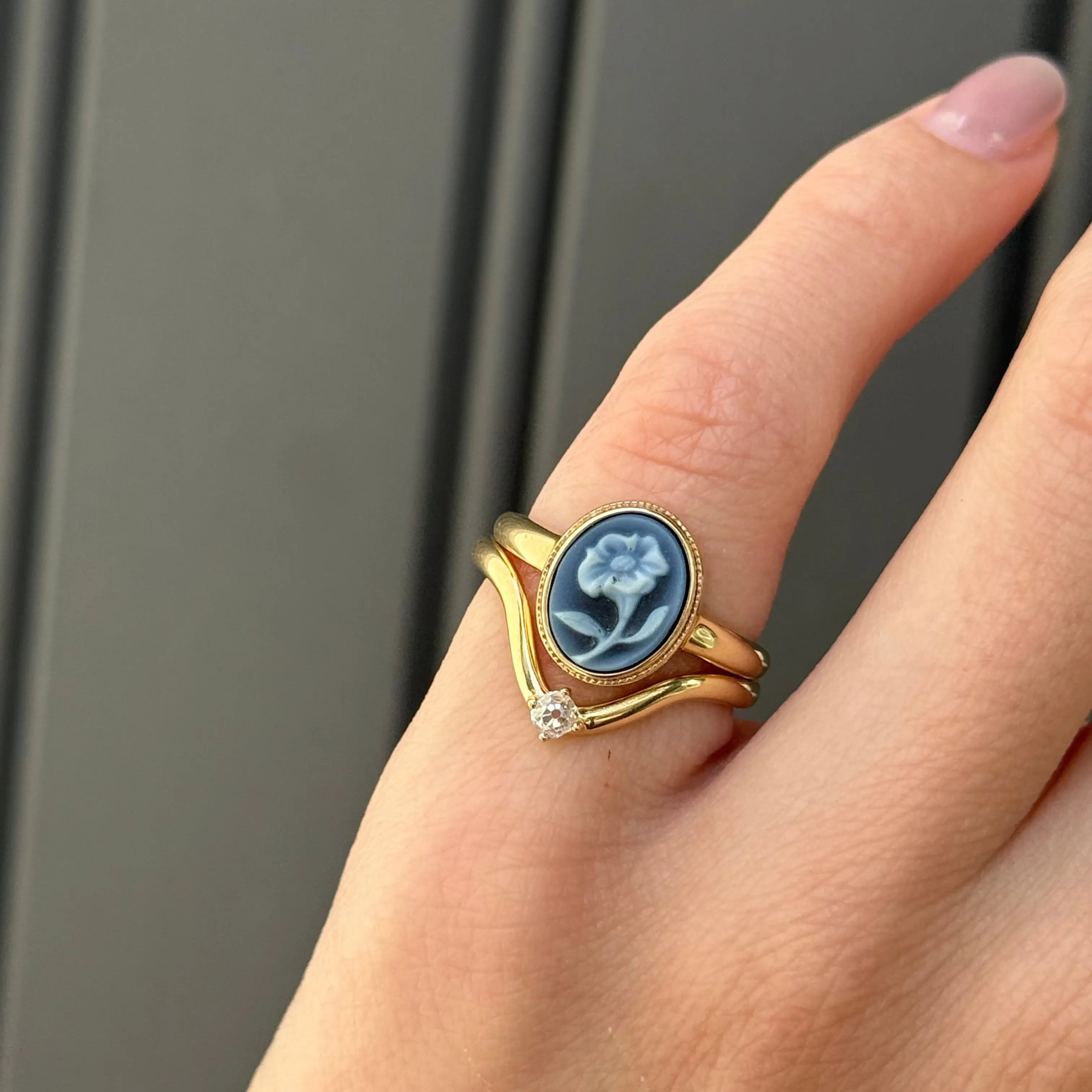 Photo of two rings on a pinky: one gold ring with a blue flower carving and one wavy gold stacking ring with a small diamond. Shown against a black background.