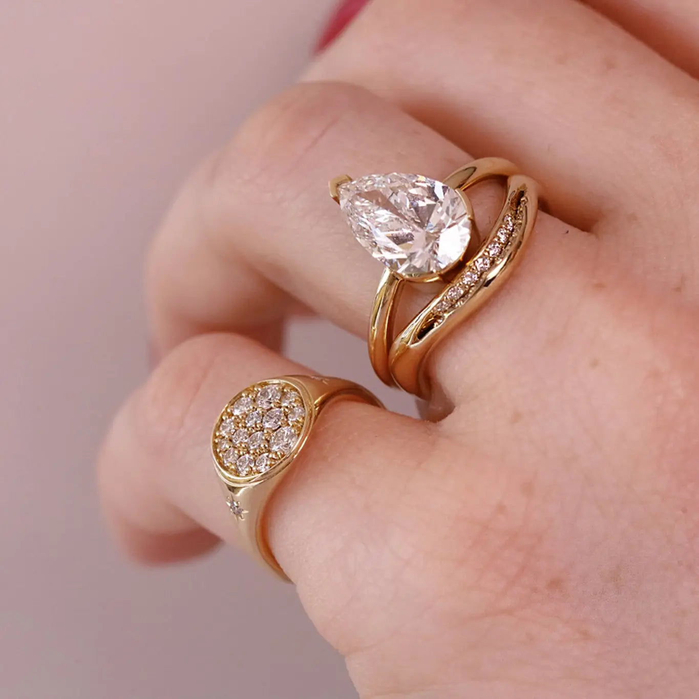 A close-up of a hand wearing two gold rings. The larger ring features a pear-cut clear gemstone, and the smaller ring has a round pavé-set clear gemstone, both on a soft pink background.